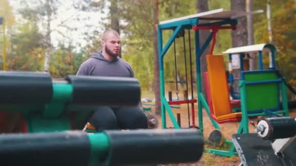 Чоловік бодібілдер піднімає гантелі тренування на відкритому повітрі дитячий спортивний майданчик — стокове відео