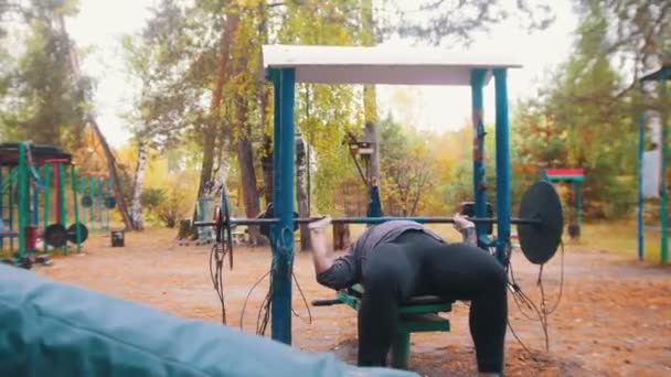 Un culturista hombre tatuado levanta una enorme mancuerna pesada - entrenamiento en el campo de deportes para niños al aire libre — Vídeo de stock