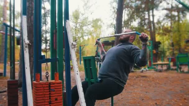 屋外のスポーツグラウンドで男性ボディービルダーのトレーニング - 重量に接続されたバーを引っ張る - 秋の森 — ストック動画