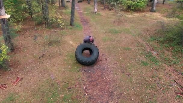 A tough man bodybuilder pushes over the tire on the ground in the forest — Stock Video