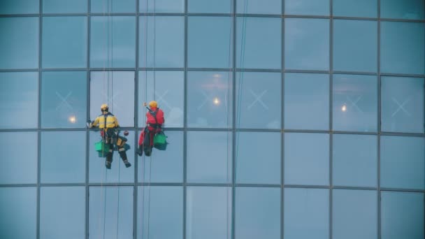 Двое рабочих в красной и желтой рабочей одежде чистят наружные окна небоскреба - индустриальный альпинизм — стоковое видео