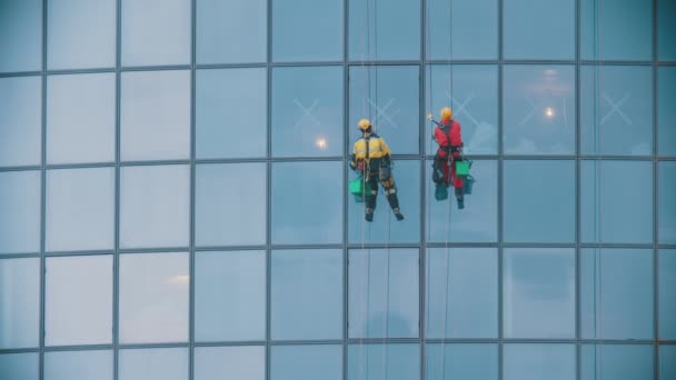 Twee mannen in rood en geel werk kleding schoonmaken van de buitenkant blauwe ramen van een wolkenkrabber - industriële alpinisme — Stockvideo