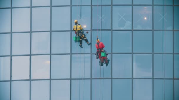 Dos hombres trabajadores en ropas de trabajo rojas y amarillas limpiando las ventanas exteriores de un rascacielos de negocios - alpinismo industrial — Vídeo de stock