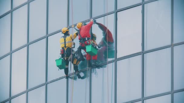 Két férfi munkás kötélen lóg egy üzleti felhőkarcoló külső kék ablakainál, és megtisztítja őket - ipari alpinizmus - ősszel és felhős időben — Stock videók