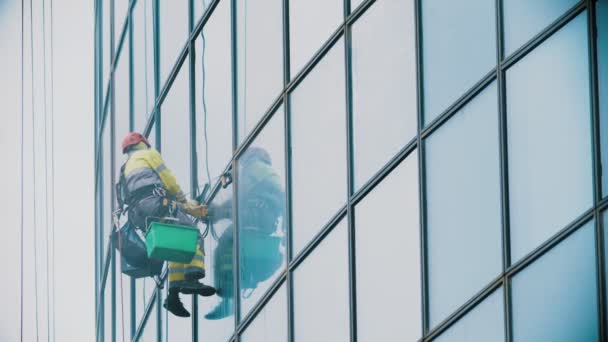 Человек-рабочий, висящий на веревках и вытирающий внешние окна бизнес-небоскреба - индустриальный альпинизм - пасмурная погода — стоковое видео