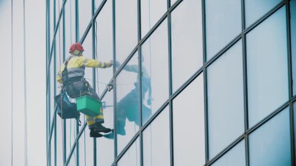 Un travailleur en vêtements de travail jaunes accrochés à des cordes et essuie les fenêtres extérieures d'un gratte-ciel d'entreprise - alpinisme industriel - temps couvert — Video