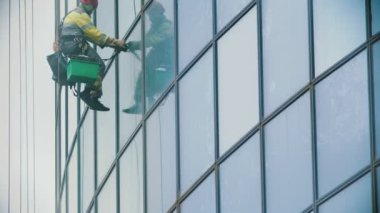 Sarı iş elbiseleri giyen bir işçi, bir iş gökdeleninin dış camlarını pislikten temizliyor. Endüstriyel dağcılık, bulutlu bir hava.
