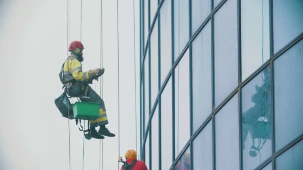Kötélen lógó munkások, akik letörlik egy üzleti felhőkarcoló külső ablakait - az üvegre szerelnek egy eszközt - ipari alpinizmus - borús időjárás — Stock videók