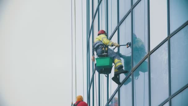 Ein Mann in gelber Arbeitskleidung hängt an Seilen und wischt die Außenfenster eines Wolkenkratzers ab - hält sich an der am Glas befestigten Vorrichtung fest - industrieller Alpinismus - bewölktes Wetter — Stockvideo