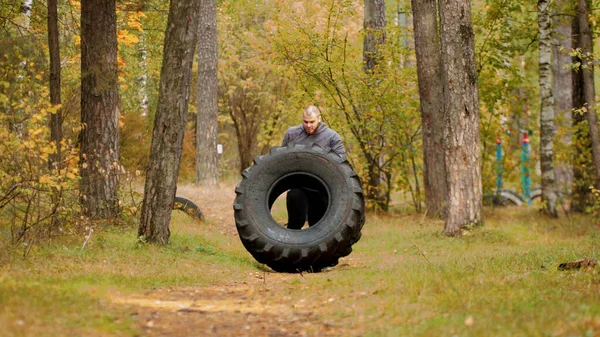 A big man bodybuilder turning over the tire on the floor and moving it - training outdoors