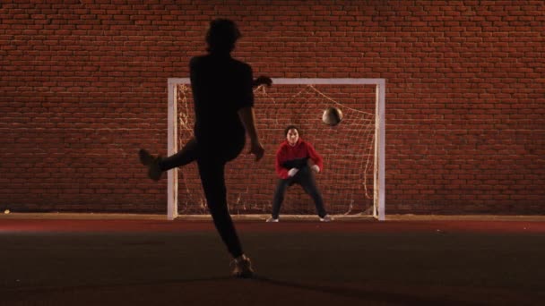 Двоє молодих друзів грають у футбол на відкритому майданчику вночі захищаючи футбольні ворота — стокове відео
