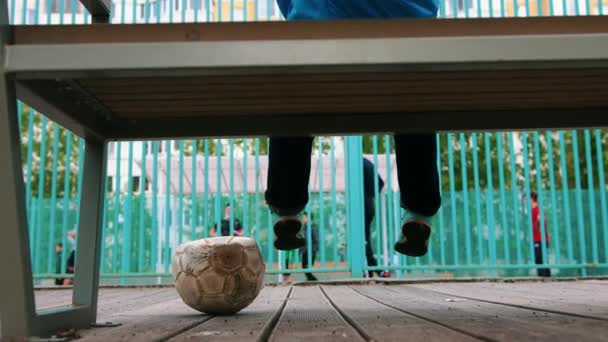 Ein kleiner Junge sitzt auf der Bank und beobachtet andere Kinder beim Fußballspielen — Stockvideo