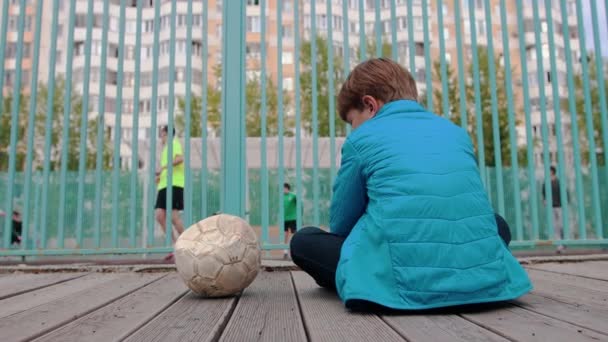 Un niño pequeño sentado en el banco cerca de la pelota desinflada y viendo a otros niños jugando al fútbol — Vídeo de stock