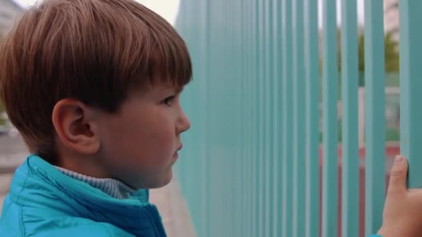 一个悲伤的小男孩看着另一个孩子在格子后面踢足球 — 图库视频影像