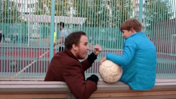 Joven hombre sonriente y su hermano pequeño sentado en el banco y viendo a otros niños jugando al fútbol — Vídeos de Stock