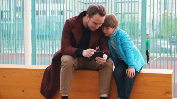 Joven hombre y su hermano pequeño sentado en el banco y mirando la pantalla del teléfono — Vídeo de stock