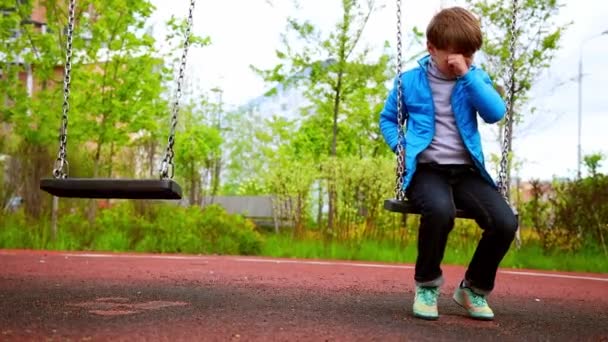 Трохи сумний самотній хлопчик сидить на гойдалках на дитячому майданчику — стокове відео