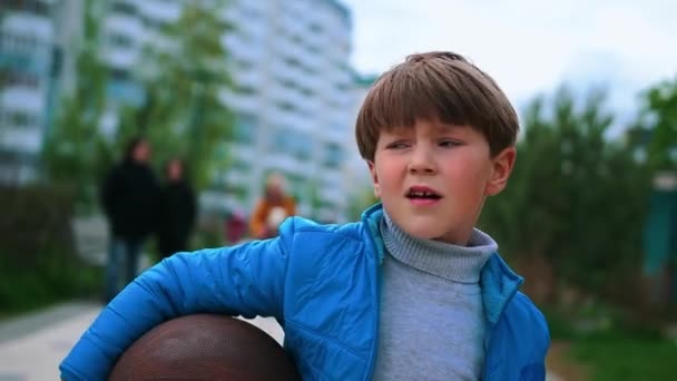 Маленький мальчик ходит по улице, держа баскетбольный мяч. — стоковое видео