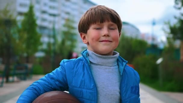 Een kleine lachende jongen die op straat loopt en basketbal vasthoudt. — Stockvideo