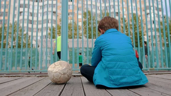 Ein kleiner Junge sitzt auf der Bank neben dem entleerten Ball und beobachtet andere Kinder beim Fußballspielen — Stockfoto