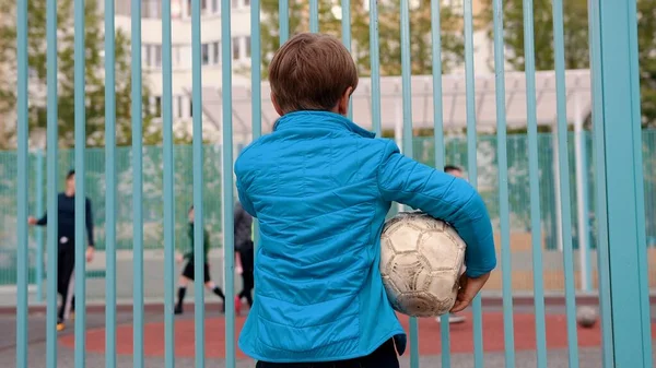 Un niño inseguro sosteniendo una pelota desinflada y viendo a otros niños jugando al fútbol —  Fotos de Stock