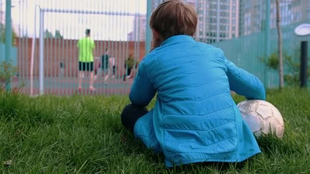 Ein kleiner Junge sitzt mit Fußballball auf dem Rasen und sieht anderen beim Spielen zu — Stockvideo