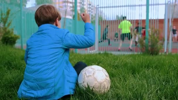サッカーボールを持って芝生の上に座って他の人が遊び場で遊んでいるのを見ている小さな男の子 — ストック動画