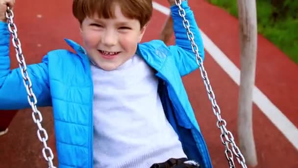 Молодой человек размахивает своим улыбающимся младшим братом на качелях на детской площадке — стоковое видео