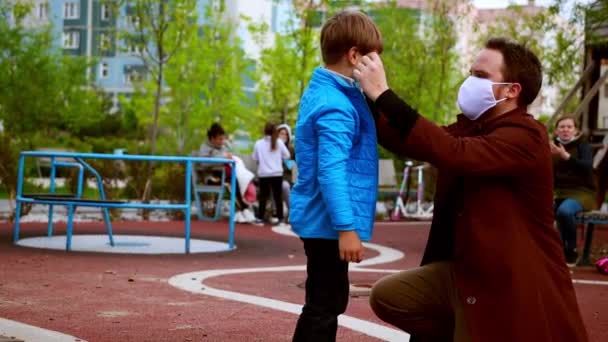 Jonge man en kleine jongen lopen op de speelplaats - zorgzame man zet een medisch masker op zijn broer — Stockvideo