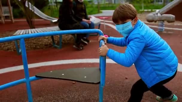 बाहेरच्या खेळाच्या मैदानावर खेळत वैद्यकीय मास्कचा एक लहान मुलगा स्विंगला गती देतो — स्टॉक व्हिडिओ