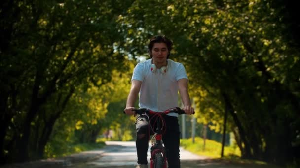 Joven hombre feliz en camisa blanca montando una bicicleta y poniéndose los auriculares — Vídeo de stock