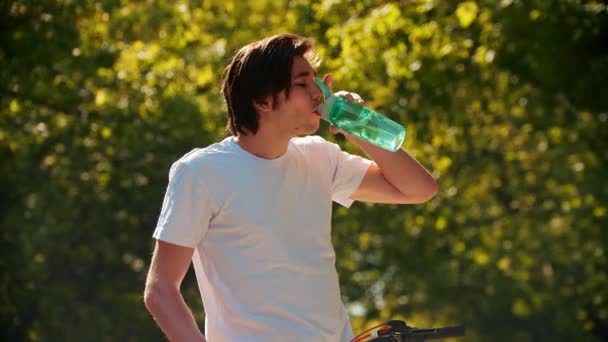 Junger Mann im weißen Hemd trinkt Wasser aus der Flasche und radelt weiter — Stockvideo