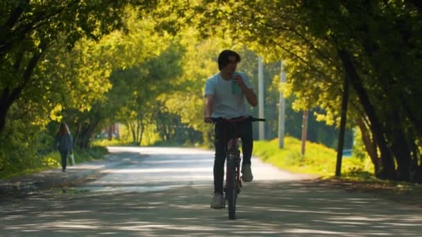 Fiatal sportos férfi fehér ingben biciklizik és vizet iszik az üvegből. — Stock videók