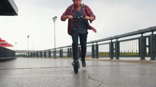 Joven hombre sonriente montando un scooter eléctrico en el muelle cerca del río — Vídeos de Stock