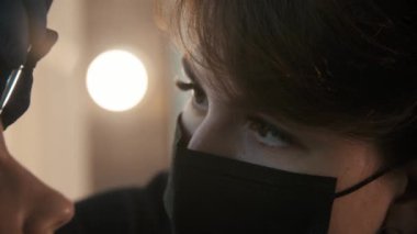 Siyah maskeli genç bir kozmetik uzmanı modelin kaşlarını koyu renklerle dolduruyor.