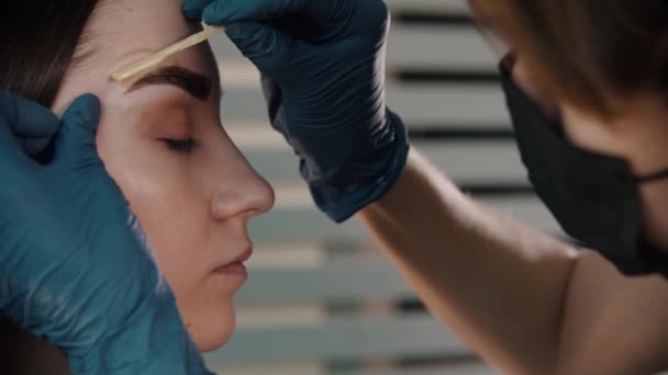 Brow master i svart mask tillämpa vax på ögonbrynen på ung kvinna modell — Stockvideo