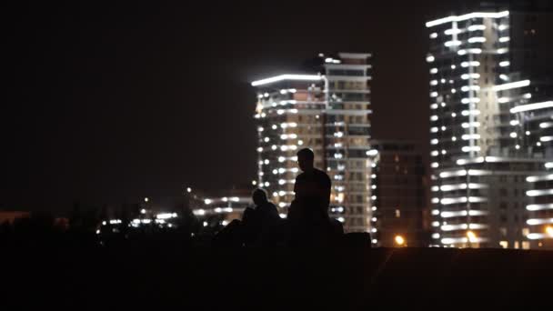 Junge Männer und Frauen sitzen nachts am Strand vor dem Hintergrund glühender Wolkenkratzer — Stockvideo