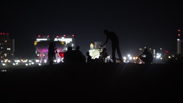 Giovani seduti sulla spiaggia di notte sullo sfondo di grattacieli incandescenti e divertirsi — Video Stock