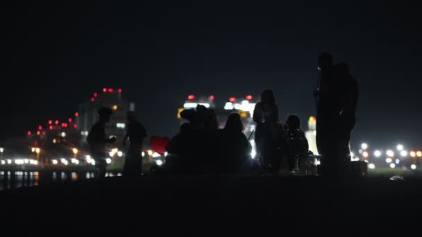 Mladí lidé sedí v noci na pláži na pozadí zářících mrakodrapů a baví - kouření hookah a tanec — Stock video