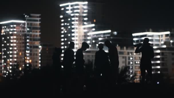 Młodzi ludzie urządzają imprezę na plaży w nocy na tle świecących wieżowców i dobrze się bawią - piją alkohol — Wideo stockowe