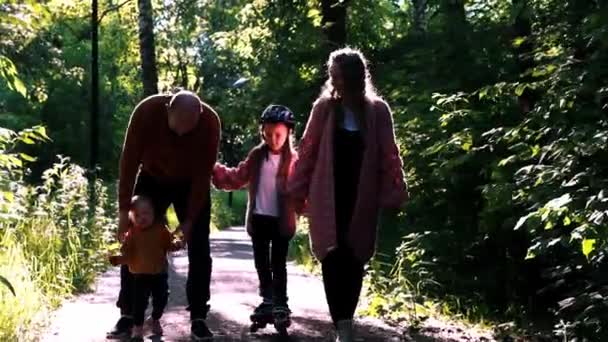 Jonge gelukkige familie van ouders en twee kleine kinderen wandelen in het park - een meisje schaatsen op de rollen — Stockvideo