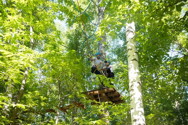 Rope adventure - woman and man having a rope entertainment in the green forest - standing on high and walking on the rope bridge