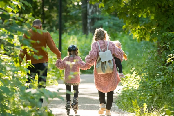 Jong gelukkig gezin van ouders en twee kleine kinderen wandelen in het bos - mam en pap houden hun dochter op skate rollen door de handen — Stockfoto