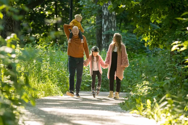 Familia joven de padres y dos niños pequeños caminando en el bosque: mamá y papá sosteniendo a su hija en patines por las manos — Foto de Stock