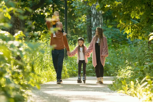 Jonge familie van ouders en twee kleine kinderen wandelen in het park - mam en pap houden hun dochter op skate rollers door de handen — Stockfoto