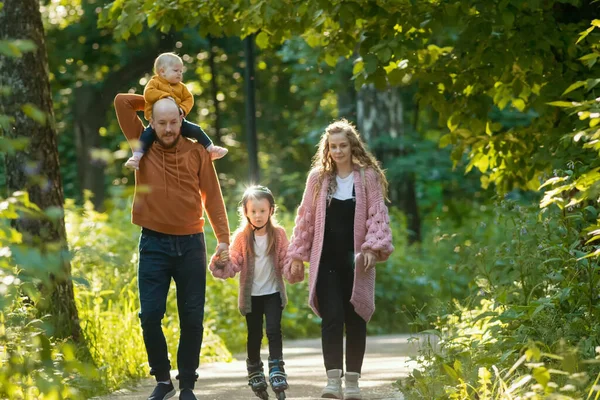 Jonge familie van ouders en twee kinderen wandelen in het park - mam en pap houden hun dochter op skate rollers door de handen — Stockfoto