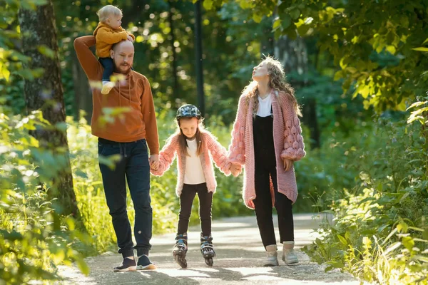 年轻快乐的父母和两个孩子在公园里散步- -爸爸妈妈牵着女儿的手滑旱冰 — 图库照片