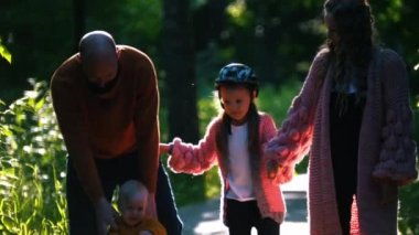 Küçük bir aile ve iki küçük çocuk yaz parkında yürüyor - küçük bir bebek yürüyor ve bir kız paten kayıyor