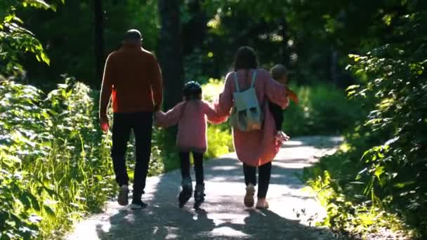 Junge Familie mit Eltern und zwei kleinen Kindern beim Spazierengehen im Sommerpark - Mama und Papa halten ihre Tochter an den Händen auf Rollen — Stockvideo