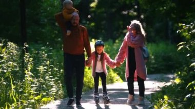 Mutlu ebeveynler ve iki küçük çocuk yaz parkında yürüyorlar. Anne ve baba, kızlarını el yordamıyla tutuyorlar.